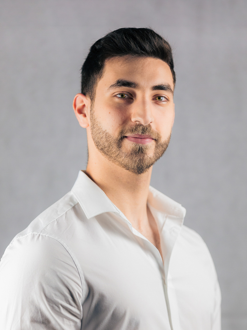 A headshot of Mohammed Saffouri, who looks at the camera from a slight angle. He has dark hair and a short beard, smiling at the camera. He wears a white button down shirt and is pictured in front of a neutral gray background.