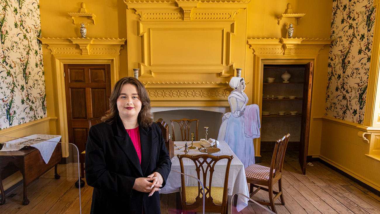 Katherine Ashby standing in the Gunston Hall mansion/museum and looking at the camera. Behind her in the exhibit is one of her depictions of an enslaved woman who is holding a cloth. The depiction is placed in a room that has a table, chairs, and dining wear on the table.