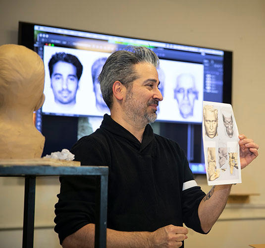 Joe Mullins holding up a piece of paper to the forensic artists sculpture class showing sketches of faces standing in front of large computer screen with more faces