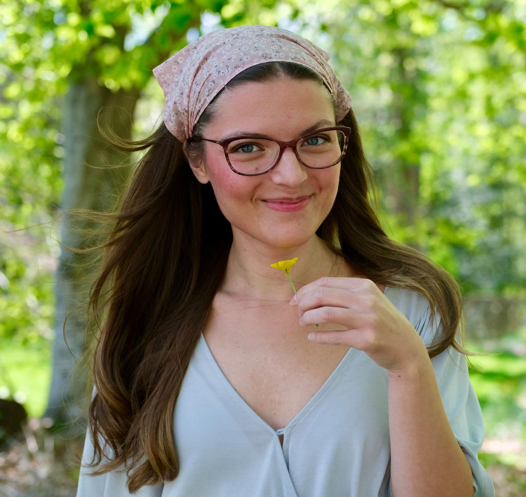 Brianna Camp art alum from School of Art - young white woman smiling, with long brown hair wearing a headscarf and glasses, a blue top and holding a yellow flower up to her face