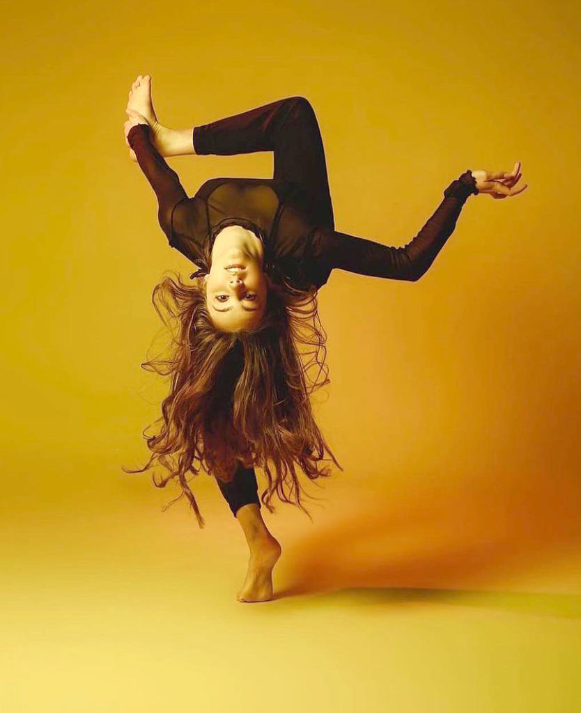 Dancer Sydney Heldman bending over backwards looking at us upside down wearing black against yellow background