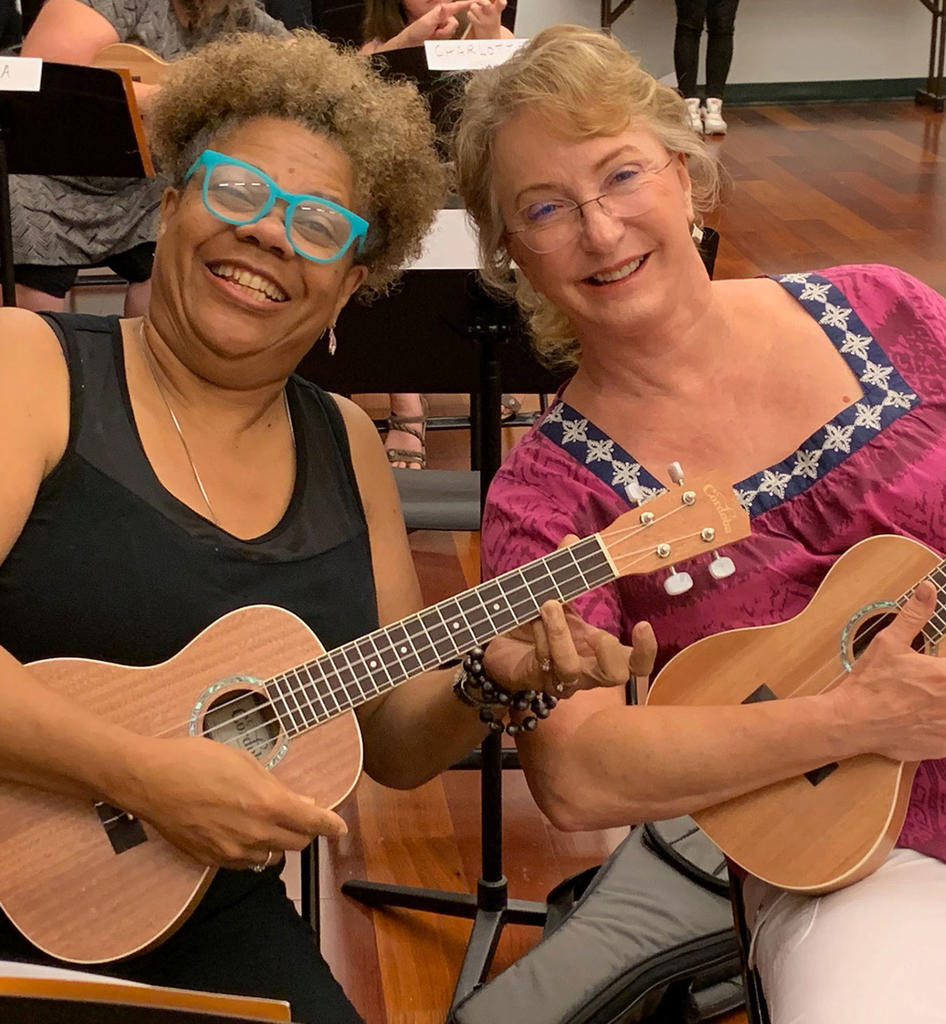 two ladies, one white and one black, sitting side by side at the Hylton Performing Arts Center with a ukelele on their laps