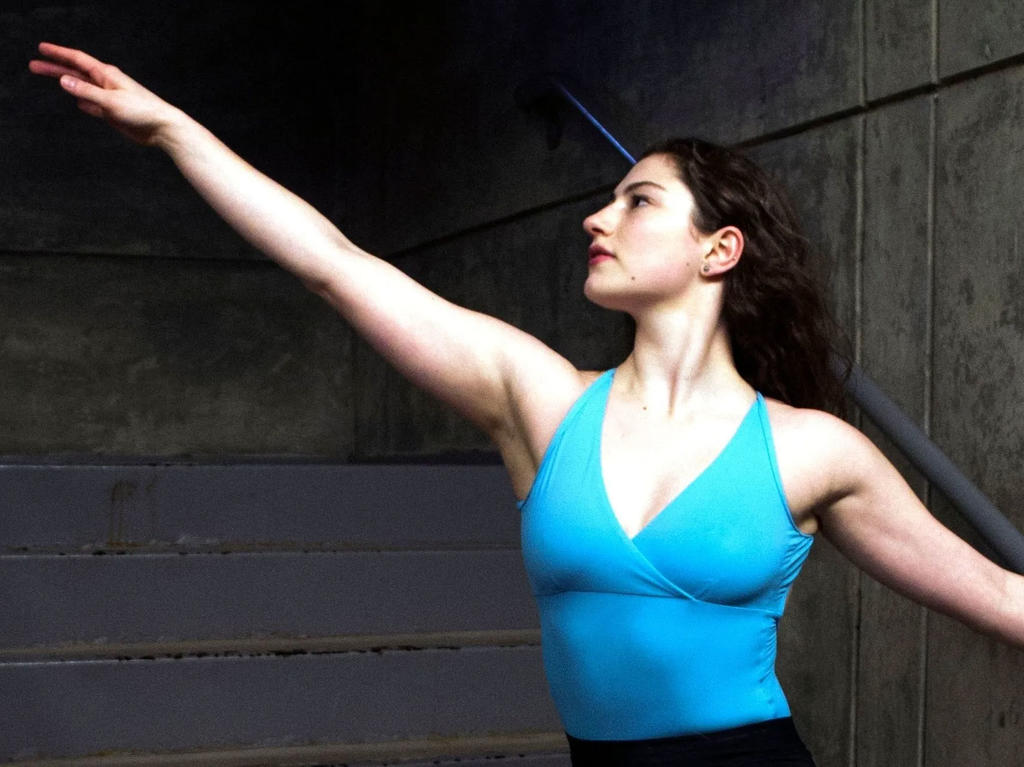 white woman with long brown hair dressed in blue sleeveless leotard turned to the side with one arm stretched upwards in front and the other behind and down
