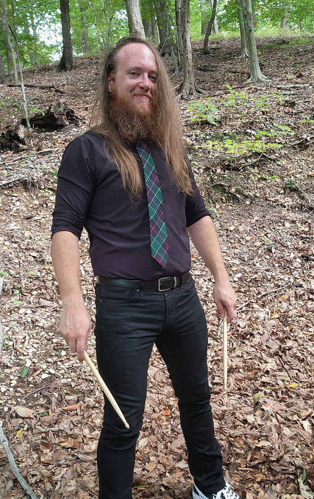 Tall thin white man with long brown hair and a bushy beard wearing a purplel shirt and plaid tie with black jeans carrying drumsticks against backdrop of woods