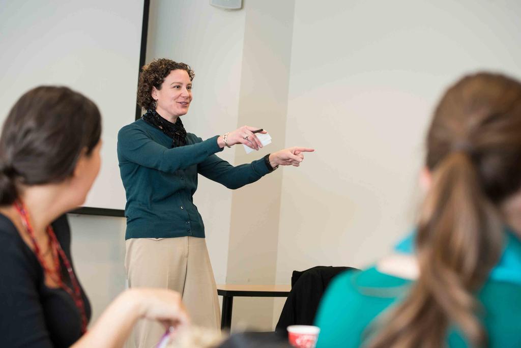 white woman with short brown curly hair wearing green cardigan and beige pants talking in front of class