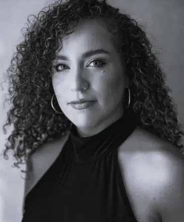 Adelina Mitchell is a young white female with crinkly shoulder length dark hair wearing a black halter necked top with bare shoulders and large hoop earrings