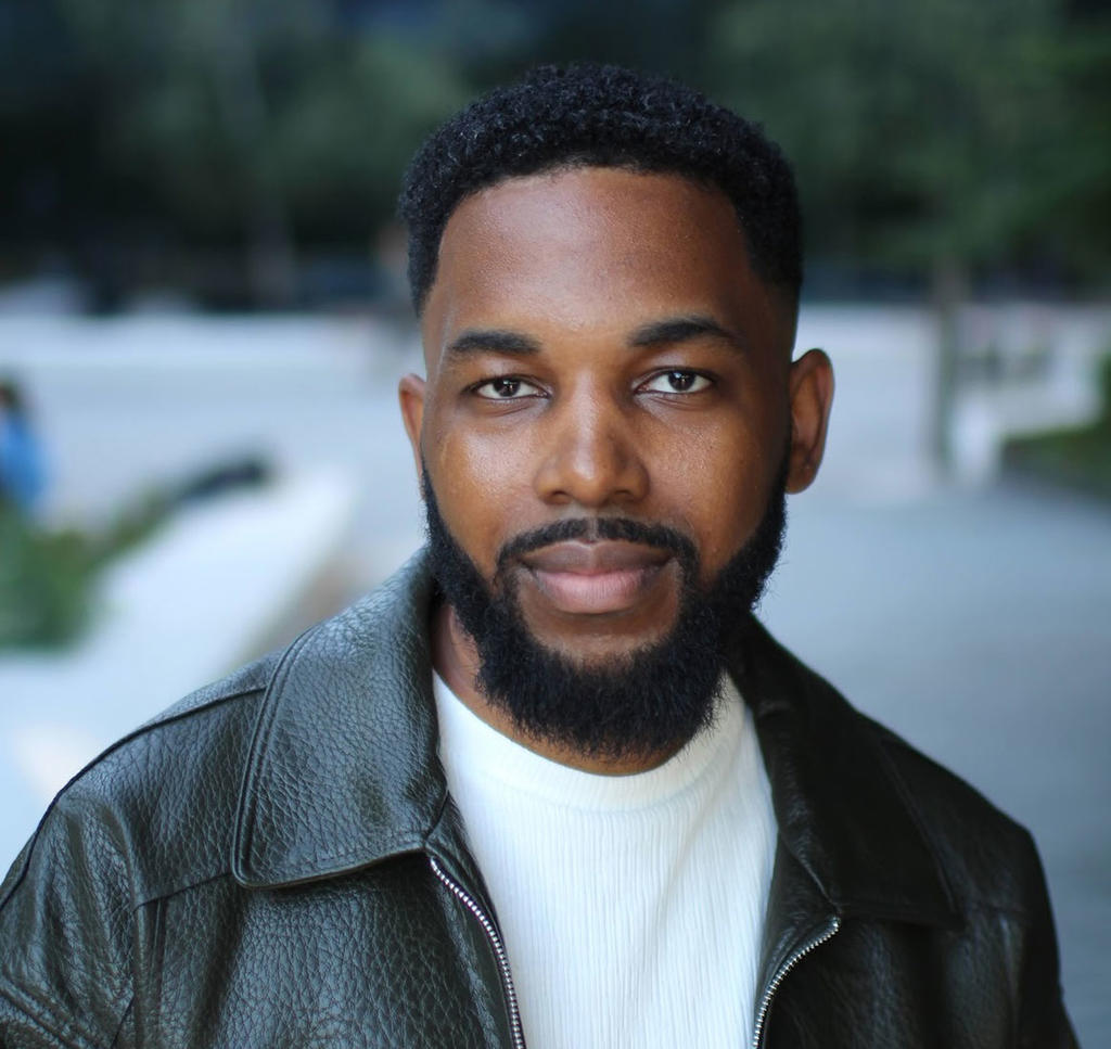 Taj Kokayi: a young black male with beard and mustache, short black hair, wearing black leather jacket and white T shirt